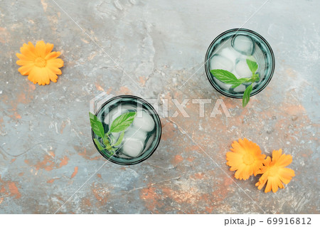 Two beautiful vintage turquoise glasses with cold drink and ice cubes, with fresh green mint leaves decorated with marigold flowers on natural colorful stone table. Top view. Two beautiful vintage turquoise glasses with cold drink and ice cubes, with fresh green mint leaves decorated with marigold flowers on natural colorful stone table. Top view. 69916812