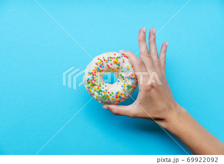 One donut with white icing. An overhead view of a doughnut with colored sprinkles. Female hand holding donut over blue background. Top view, flat lay. Sweet, diet concept. 69922092