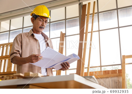 Asian old man with yellow safety helmet checking blueprint in carpentry workshop. Concept senior people doing hobby after retirement. Asian old man with yellow safety helmet checking blueprint in carpentry workshop. Concept senior people doing hobby after retirement. 69932935