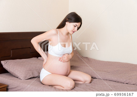 beautiful pregnant woman touching her tummy and keeping one hand on her back at home on bed 69936167