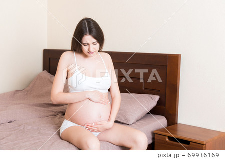 Young pretty pregnant woman, in pain, sitting on bed in bedroom, preparing to give a birth 69936169