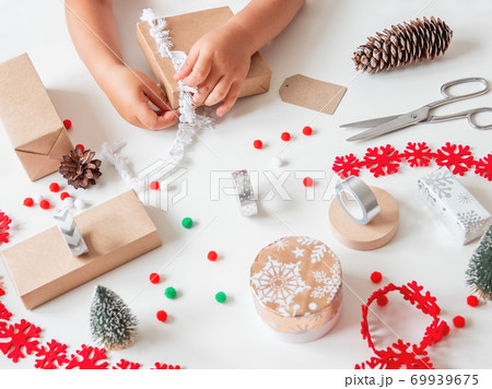 Kid wraps handmade Christmas presents in craft paper with colorful pompons and snowflake ribbons. Child prepares gifts for New Year celebration. Peaceful leisure activity before winter holidays. Kid wraps handmade Christmas presents in craft paper with colorful pompons and snowflake ribbons. Child prepares gifts for New Year celebration. Peaceful leisure activity before winter holidays. 69939675