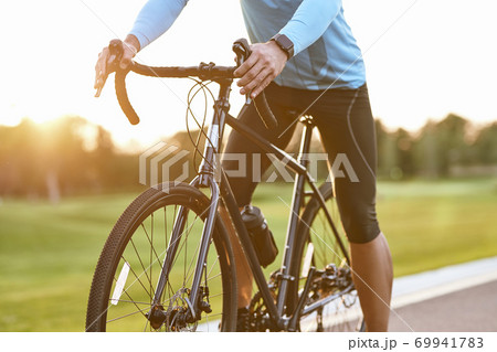 Cropped shot of professional road bicycle racer in sportswear cycling outdoors at sunset, man standing with bicycle on the road outdoors 69941783