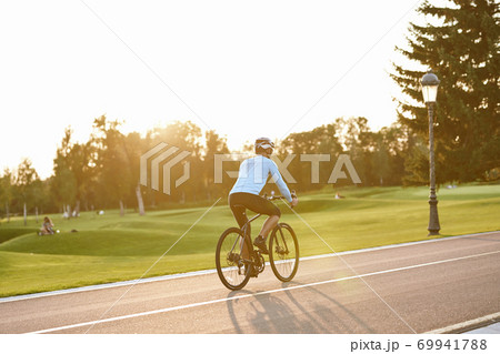 Perfect day for cycling. Rear view of athletic man in sportswear riding bicycle along the road in city park at sunset Perfect day for cycling. Rear view of athletic man in sportswear riding bicycle along the road in city park at sunset 69941788