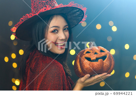 Portrait of beautiful young woman in witch halloween costume wear witches hat hold pumpkin Jack O Lanterns over spooky dark magic background - Halloween party concept Portrait of beautiful young woman in witch halloween costume wear witches hat hold pumpkin Jack O Lanterns over spooky dark magic background - Halloween party concept 69944221