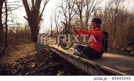 Happy caucasian woman cyclist makes a photo of herself a selfie on the phone while sitting on the bridge over a river in the forest in sunny weather 69945649