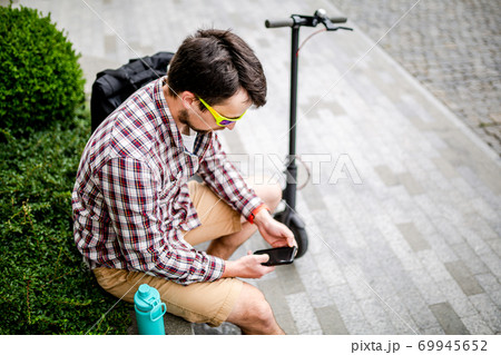 Individual urban environmental friendly transport electric kick scooter parked near man sitting on wooden bench in recreation area. Male use phone, surfing Internet, online shopping at smartphone 69945652