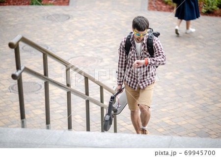 Man walking with folded electric scooter up stairs in city near modern building. Ecological technological lifestyle. E-Mobility. Person Carrying Electric Scooter Folded Position. Eco urban transport Man walking with folded electric scooter up stairs in city near modern building. Ecological technological lifestyle. E-Mobility. Person Carrying Electric Scooter Folded Position. Eco urban transport 69945700
