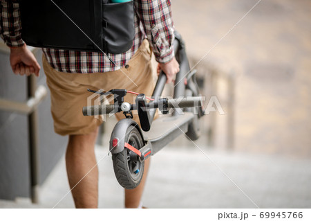 Man Carrying folded E-Scooter in hand on stairs near building. Moving by eco urban transport, modern city concept. Ecological technological lifestyle. Person Carry Electric Scooter In Folded Position Man Carrying folded E-Scooter in hand on stairs near building. Moving by eco urban transport, modern city concept. Ecological technological lifestyle. Person Carry Electric Scooter In Folded Position 69945766