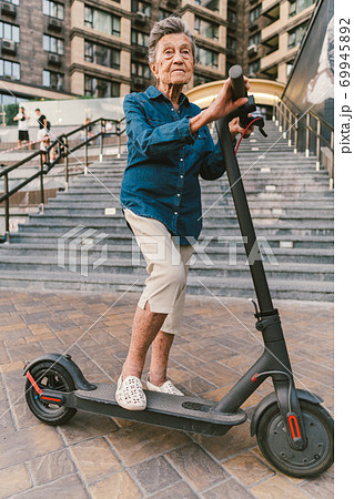 Cheerful elderly woman on a scooter. Grandma rides a scooter. Grandmother using urban personal transporter. Active retirement concept. Healthy cheerful senior retired lady. Ecological transport Cheerful elderly woman on a scooter. Grandma rides a scooter. Grandmother using urban personal transporter. Active retirement concept. Healthy cheerful senior retired lady. Ecological transport 69945892