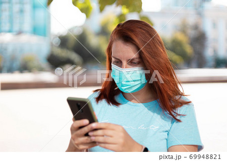Portrait of a young beautiful girl in a medical protective mask looks and uses a smartphone in a park on the street 69948821