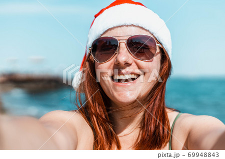 Beautiful happy girl in santa hat and swimsuit makes selfie and enjoying winter holidays getaway on tropical beach. Beautiful happy girl in santa hat and swimsuit makes selfie and enjoying winter holidays getaway on tropical beach. 69948843