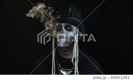 Sinister man with horrible Halloween holiday skeleton makeup raising his head and smoking cigar 69951261