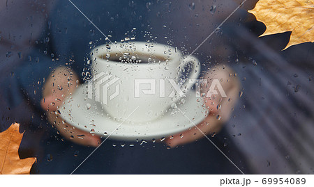 Woman hands holding cup of coffee through window with raindrops Woman hands holding cup of coffee through window with raindrops 69954089