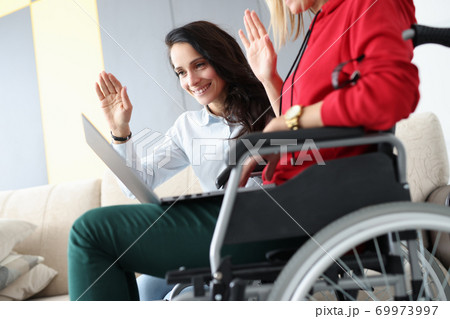 Woman in wheelchair with her friend communicate via internet laptop waving hand Woman in wheelchair with her friend communicate via internet laptop waving hand 69973997