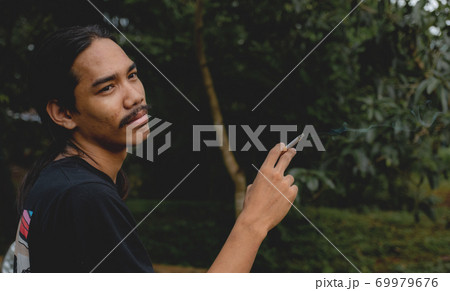 A long-haired Asian man with a beard holding a cigarette standing outside A long-haired Asian man with a beard holding a cigarette standing outside 69979676