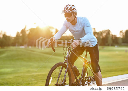 Professional road bicycle racer in sportswear and protective helmet standing on the road at sunset, ready to ride. Man cycling in the park Professional road bicycle racer in sportswear and protective helmet standing on the road at sunset, ready to ride. Man cycling in the park 69985742