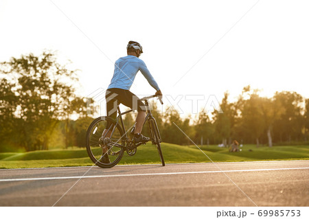 Rear view of athletic man in sportswear cycling outdoors at sunset, professional racer standing with bicycle on the road and enjoying amazing nature view Rear view of athletic man in sportswear cycling outdoors at sunset, professional racer standing with bicycle on the road and enjoying amazing nature view 69985753