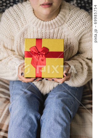 Young woman sits on bed in cozy knitted wool white sweater and holding gift-box with red gold. Hygge, New Years, Christmas, holiday preparations. Candles, Christmas balls, gifts, cocoa marshmallows. Young woman sits on bed in cozy knitted wool white sweater and holding gift-box with red gold. Hygge, New Years, Christmas, holiday preparations. Candles, Christmas balls, gifts, cocoa marshmallows. 69985754