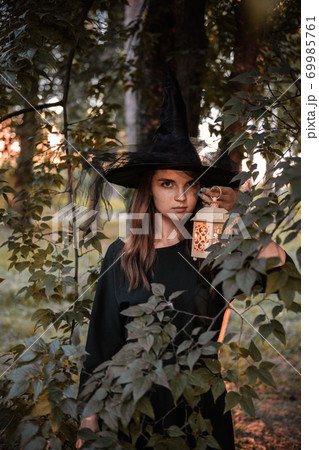 Young woman in dark dress and witch's hat holds lantern with candles in her hands and illuminates the forest. Halloween party costume. Park with autumn trees. 69985761