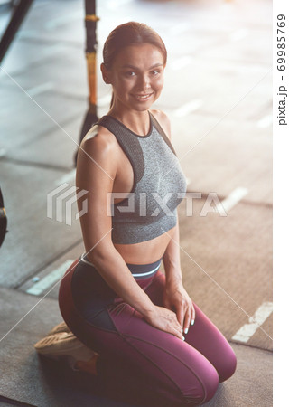 Vertical shot of a beautiful young athletic woman in sportswear kneeling down on the floor at gym, looking at camera and smiling Vertical shot of a beautiful young athletic woman in sportswear kneeling down on the floor at gym, looking at camera and smiling 69985769