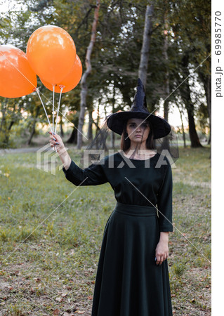 Young cute beautiful woman in dark dress and witch's hat holds orange balloons in her hands. Halloween party costume. Forest, park with autumn trees. Young cute beautiful woman in dark dress and witch's hat holds orange balloons in her hands. Halloween party costume. Forest, park with autumn trees. 69985770