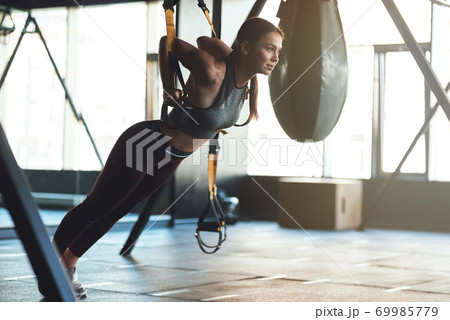 Young beautiful athletic woman wearing sports clothes exercising with fitness trx straps, doing push ups while training at gym 69985779