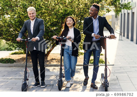 Confident happy multiracial office colleagues discussing business project go near office building with electric scooters Confident happy multiracial office colleagues discussing business project go near office building with electric scooters 69986702