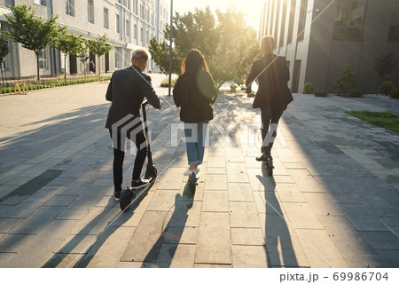 Back view of joyful international friends holding electric scooters going near modern building and talking. 69986704