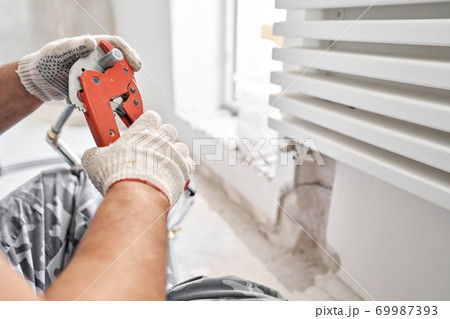 Plumber cuts the desired length of pipe with scissors tool. connection heating pipes to white Radiator in a new apartment under construction. Plumber cuts the desired length of pipe with scissors tool. connection heating pipes to white Radiator in a new apartment under construction. 69987393