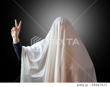 A man covered with a sheet like a ghost shows two fingers indicating victory on a dark background with backlight in the background 69987672
