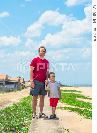 Father son walk along narrow concrete path, sea blue clear sky green grass background. Symbol as dad leads child into future adulthood. Happy childhood, fathers day, daddy influence on boy worldview Father son walk along narrow concrete path, sea blue clear sky green grass background. Symbol as dad leads child into future adulthood. Happy childhood, fathers day, daddy influence on boy worldview 69989225