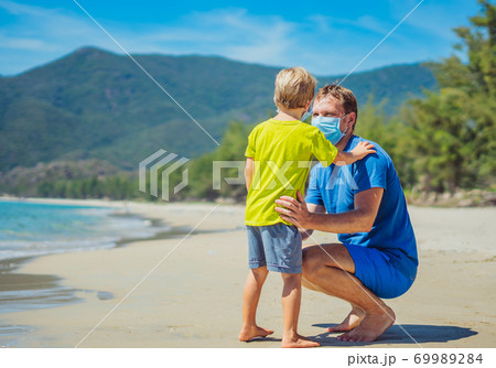 New normal reality safety during COVID 19 Global pandemic. Father and son looking on each other, wearing face blue mask protect coronavirus even walking nature sand sea beach near forest park sun day 69989284