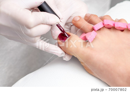 Pedicurist painting red nail polish on female toenail Pedicurist painting red nail polish on female toenail 70007802