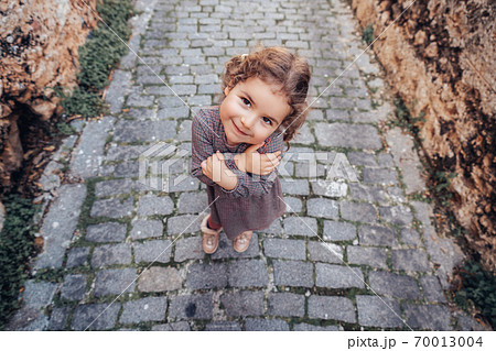Pretty baby girl hold arms closed with a shy gesture and walk on a footpath on a medieval alley. 70013004