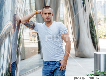 A young man leans on a shiny column. Courageous guy with a firefighter tattoo on his biceps. Male portrait. 70016759