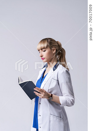 Confident young woman doctor standing with notebook or medical book in her hands. Cheks records, recipes or prescriptions. Intern in white medical clothes posing on light grey background. Confident young woman doctor standing with notebook or medical book in her hands. Cheks records, recipes or prescriptions. Intern in white medical clothes posing on light grey background. 70017009