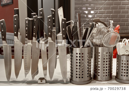 A commercial kitchen worktop, rows of knifes on a magnetic strip and utensils in pots. 70029738