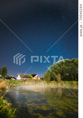Evaporation Over River Lake Near Houses In Village. Milky Way Galaxy In Night Starry Sky Above Lake River Landscape At Night. Evaporation Over River Lake Near Houses In Village. Milky Way Galaxy In Night Starry Sky Above Lake River Landscape At Night. 70040264