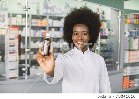 Confident smiling African woman, medical physician doctor or pharmacist posing over hospital pharmacy background, holding bottle of medicine in hands. Focus on face 70041016