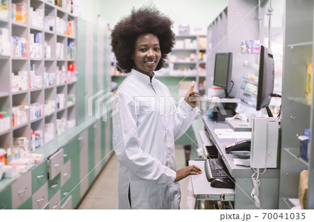 African-american woman pharmacist on her workplace in modern drugstore, standing near the counter with computer and showing thumb up. Shelves with medicines on the background African-american woman pharmacist on her workplace in modern drugstore, standing near the counter with computer and showing thumb up. Shelves with medicines on the background 70041035