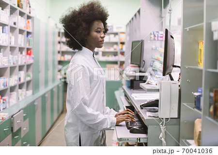Side view portrait of young confident concentrated African female pharmacist, working with computer behind counter in pharmacy Side view portrait of young confident concentrated African female pharmacist, working with computer behind counter in pharmacy 70041036