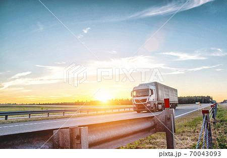 A tilt euro truck carries cargo along the highway in the evening against the backdrop of sunset. Bump stop on the road, safety, international transportation A tilt euro truck carries cargo along the highway in the evening against the backdrop of sunset. Bump stop on the road, safety, international transportation 70043093