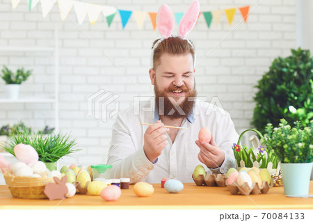 Happy easter.A funny fat man decorates eggs while sitting at a table with Easter decor in the background 70084133