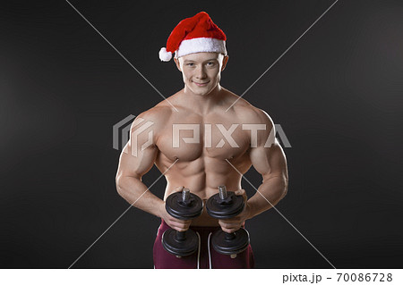 Muscular man in Santa hat with dubmbbells over dark background. Muscular man in Santa hat with dubmbbells over dark background. 70086728