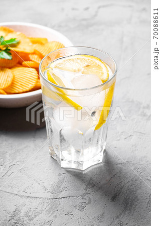 Fresh ice cold water drink with lemon near to fried corrugated golden potato chips with parsley leaf in wooden bowl on concrete background, angle view 70088611