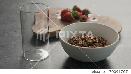 white bowl with chocolate granola, strawberries and glass for yogurt on a concrete countertop 70091767
