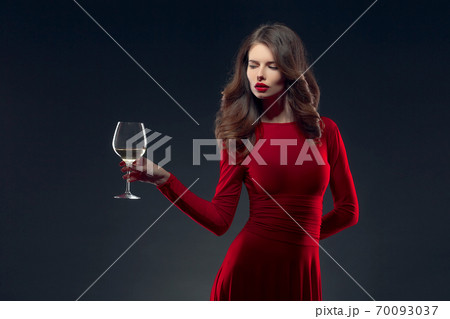 Woman with bright makeup, hairstyle wearing red dress posing with glass of vine over dark background, isolate Woman with bright makeup, hairstyle wearing red dress posing with glass of vine over dark background, isolate 70093037