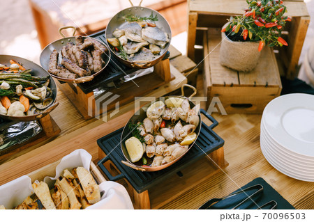 Hot pans with cooked food on a cast-iron stove on a wooden table with a bush of bitter pepper in a pot. Hot pans with cooked food on a cast-iron stove on a wooden table with a bush of bitter pepper in a pot. 70096503