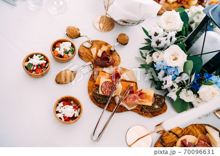 Cutting board with delicacies, kitchen tongs and spoons with a curved handle on a table with a candle in a candlestick lantern in a bouquet of flowers. Cutting board with delicacies, kitchen tongs and spoons with a curved handle on a table with a candle in a candlestick lantern in a bouquet of flowers. 70096631